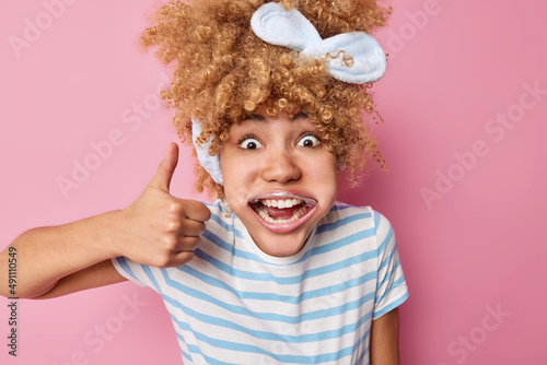 Young pretty woman uses mouth expander takes care of mouth cavity shows white teeth keeps thumb up makes okay gesture wears headband and casual striped t shirt isolated over pink background.