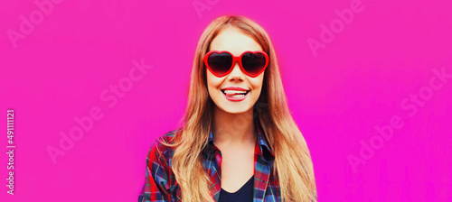 Close up portrait of happy smiling young woman teasing wearing red heart shaped sunglasses on pink background