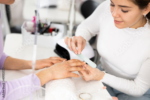 Manicure master, working in the beauty salon shaping the nails of a young female client with a nail file