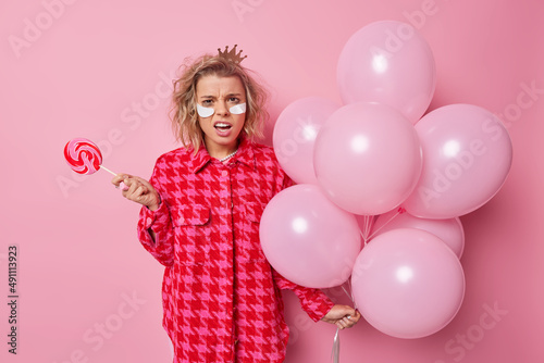 Upset displeased young woman frowns face looks indignant applies beauty white patches holds caramel candy and bunch of helium balloons wears fashionable clothes isolated over pink studio wall
