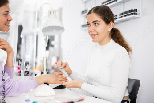 Skilled young woman manicurist filing and shaping nails of female client during procedure of manicure in beauty salon ..
