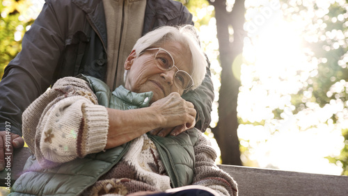 Calm elderly lady hugs her retired husband's hand while sitting on a bench in a park. High quality photo