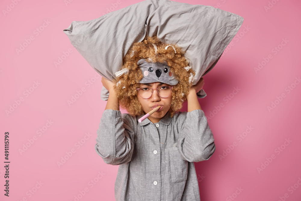 Fototapeta premium Dissatisfied curly haired woman keeps pillow over head wears sleepmask and pajama prepares for sleep isolated over pink background keeps wooden toothbrush in mouth. Sleeping rest and bedtime