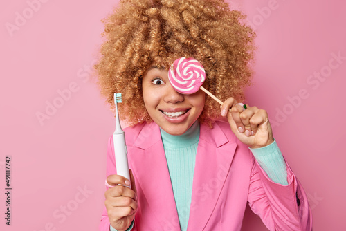 Photography Positive curly haired woman covers eye with delicious candy holds electric toothbrush has sweet tooth dressed in elegant clothes isolated over pink background