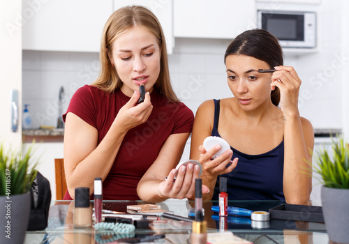 Two modern female friends having conversation and applying cosmetics in kitchen