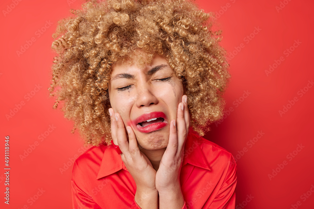 Doleful dejected curly haired woman keeps hands on cheeks cries from despair has troubles leaked makeup wears shirt isolated over vivid red background. Negative emotions and feelings concept Doleful dejected curly haired woman keeps hands on cheeks cries from despair has troubles leaked makeup wears shirt isolated over vivid red background. Negative emotions and feelings concept