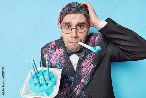 Indoor shot of puzzled brunet man dressed in festive suit blows party horn holds cake with candles celebrates birthday isolated over blue background. People celebrationn and partying concept photo