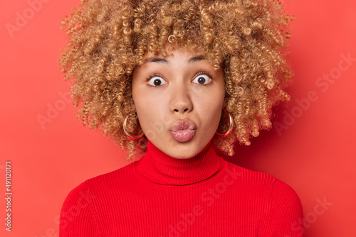 Shocked young emotional young woman has widely opened eyes keeps lips rounded sends air kiss at camera wears casual turtleneck poses against vivid red background. Human face expressions concept