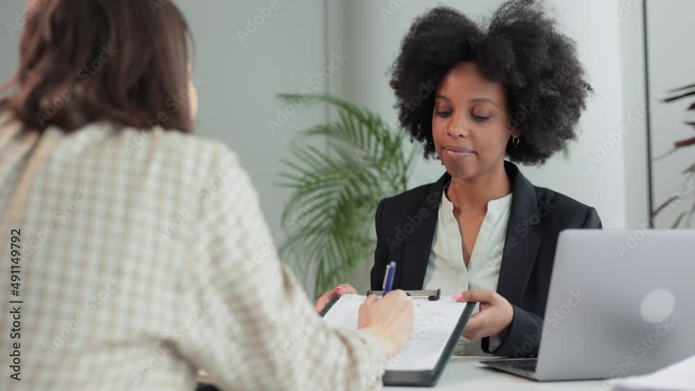 Sales manager woman, client making deal and shaking hands, sitting at table in company spbi. 4k video Young female client signs paper document and sits at desk, employee or lawyer looks with smile and