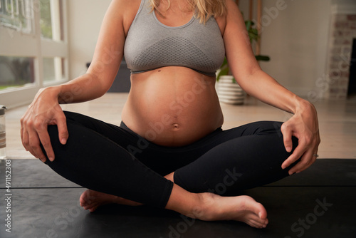 Pregnant Caucasian female sitting cross legged on yoga mat while holding her knees. Healthy lifestye in modern home. photo