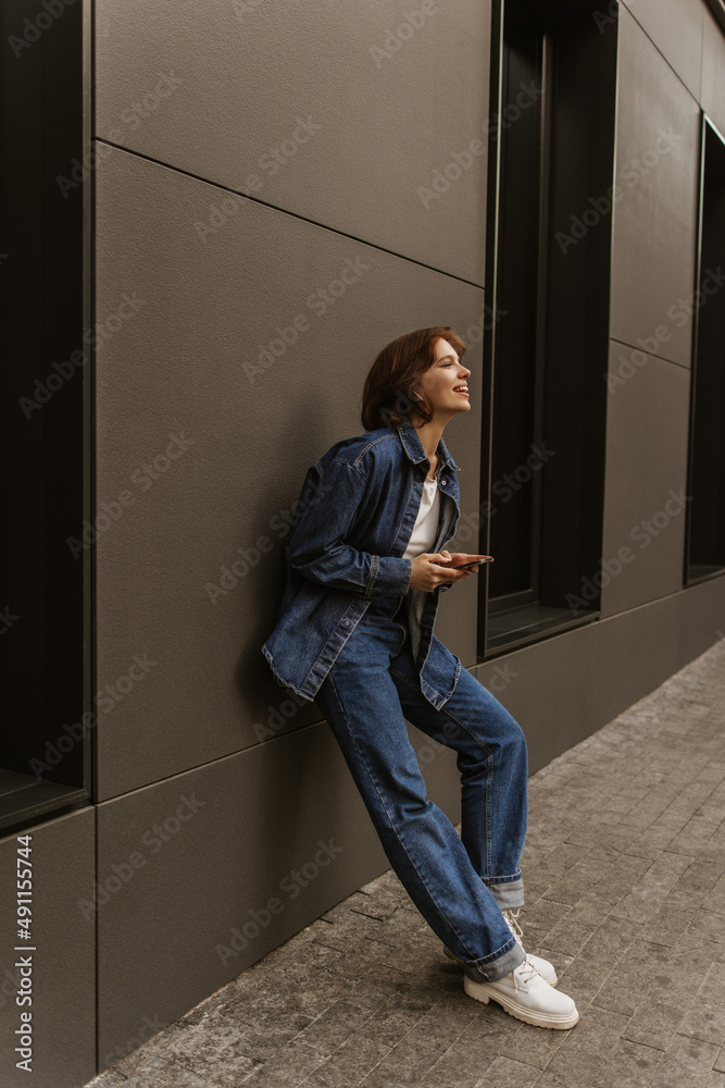 Full length image of radiant young caucasian girl standing with her back against street wall. Brunette woman is holding phone and is wearing denim clothes. Autumn trendy style
