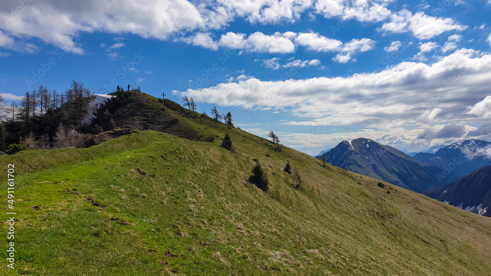 Obraz premium Hiker man at the summit cross of Rosenkogel (Rozca) with scenic view on mountain peaks in the Karawanks, Carinthia, Austria. Border with Slovenia. Triglav National Park. Alps in spring. Goal