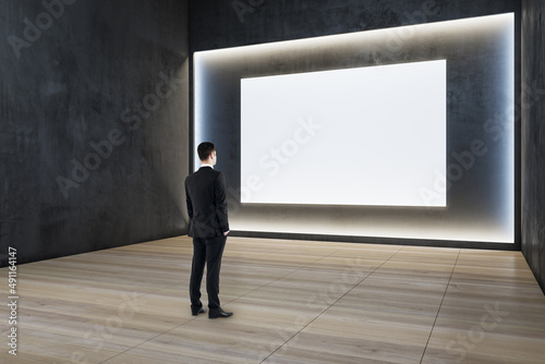 Man standing in modern exhibition hall interior with illuminated white mock up wall and wooden flooring. Gallery concept.