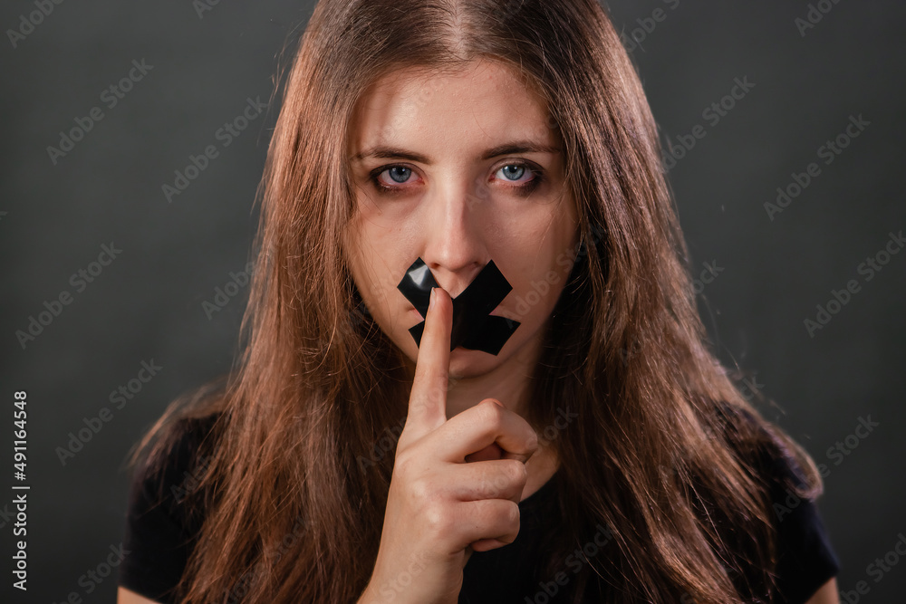 Obraz premium Portrait of a young woman with her mouth sealed with black tape, restriction of freedom of speech, censorship and a ban on speaking and expressing her opinion, isolated on a dark background.