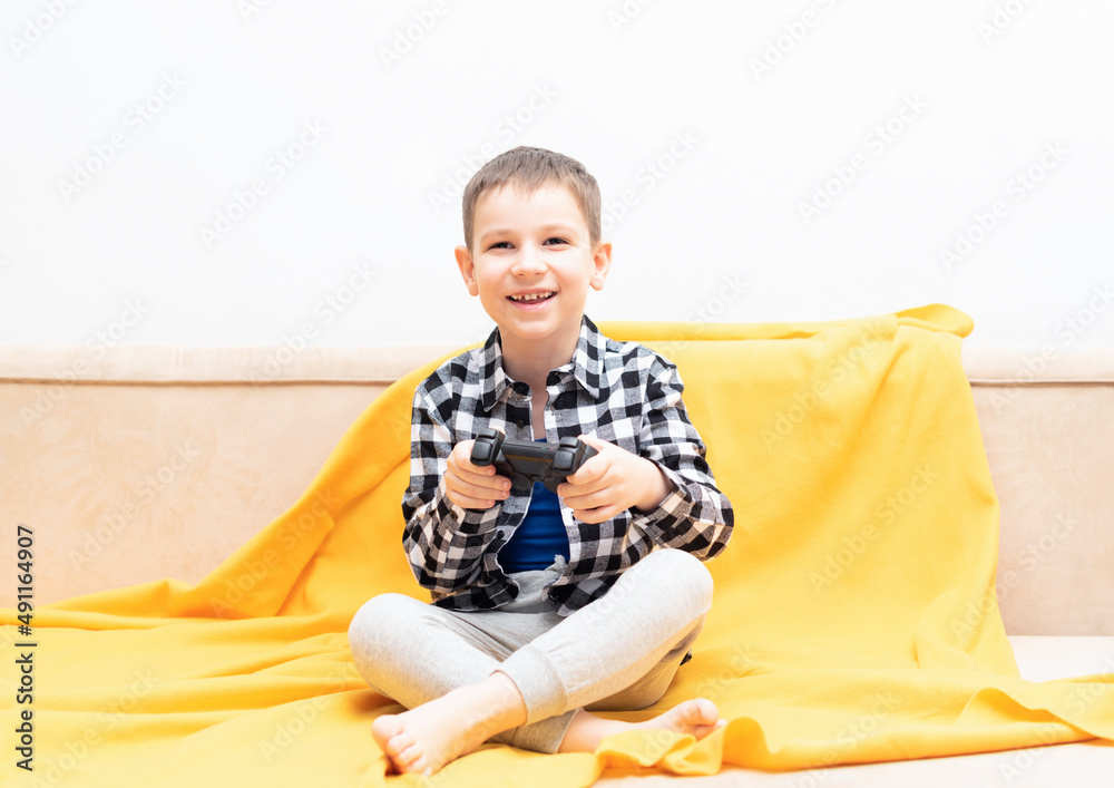 Happy child boy in checked shirt sitting on the couch with black joystick in his hands playing the video game. Playing video games at home concept Happy child boy in checked shirt sitting on the couch with black joystick in his hands playing the video game. Playing video games at home concept