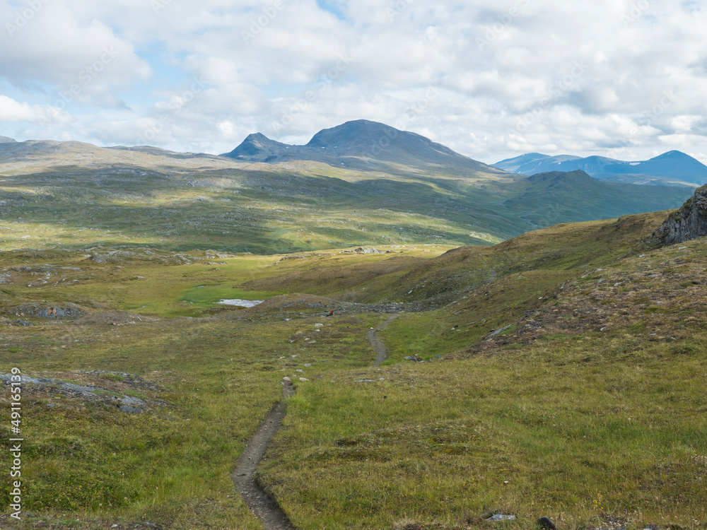 Footpath in northern artic landscape, tundra in Swedish Lapland with green hills and mountains at Padjelantaleden hiking trail. Summer day, blue sky, white clouds