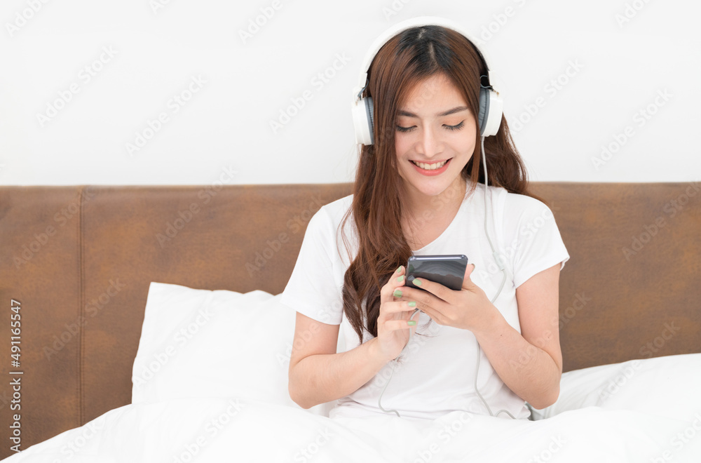 Beautiful young asian woman with headphones relaxing on the bed. She is listening to music using smartphone or tablet. Chill out and leisure concept	
