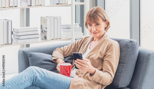 Happy beautiful relaxed mature older woman drinking coffee relaxing on sofa at living room. Elderly woman lying on sofa using smartphone and drinking coffee at home. 