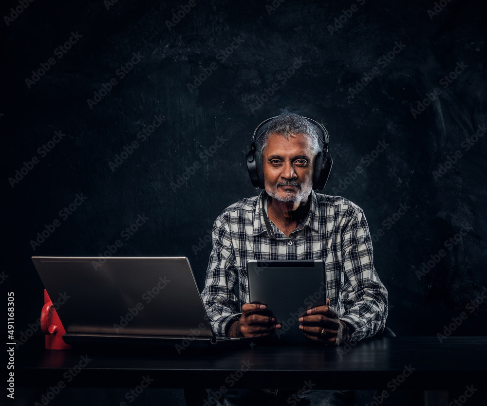 Modern old man with laptop sitting at table against dark background Modern old man with laptop sitting at table against dark background
