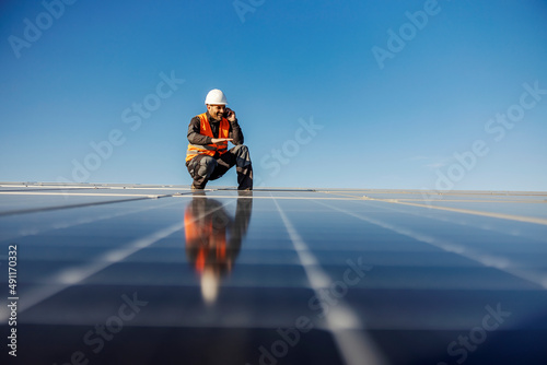 A handyman talking on the phone charged by solar power while crouching surrounded solar panels.