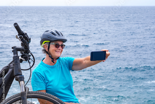 Senior ciclyst woman in outdoor excursion at sea stops on the cliff using mobile phone. Retired people with sport helmet sitting close to her bicycle holding cellphone enjoying vacation photo