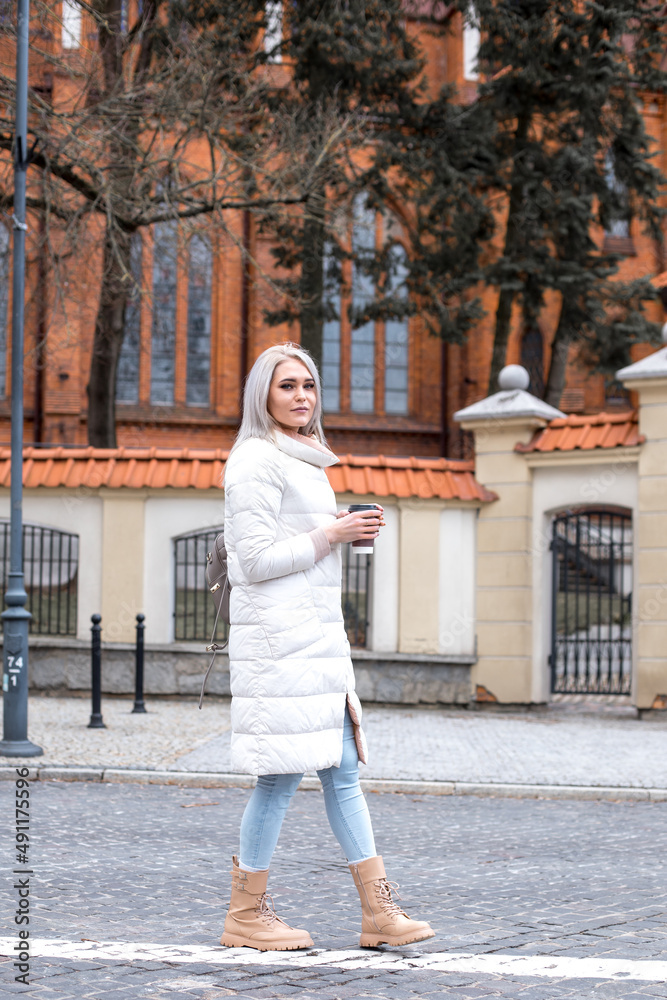 Vertical dreaming blond woman, walking down stone road alley, fashion trendy white jacket, drinking coffee, full length.