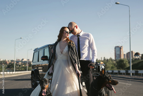 Brutal couple, bride and groom, in black glasses and leather jackets with Dobermans near the car on the road