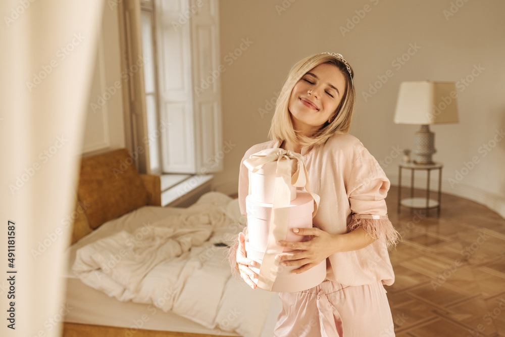 Affectionate young caucasian woman smiles tenderly, closing her eyes, holding gifts in living room. Blonde greets morning pleasantly, standing in her pajamas. Concept of seizing the moment Affectionate young caucasian woman smiles tenderly, closing her eyes, holding gifts in living room. Blonde greets morning pleasantly, standing in her pajamas. Concept of seizing the moment