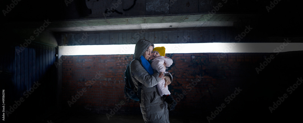 Exhausted mother holding her child in arms in the bomb shelter - obrazy, fototapety, plakaty 