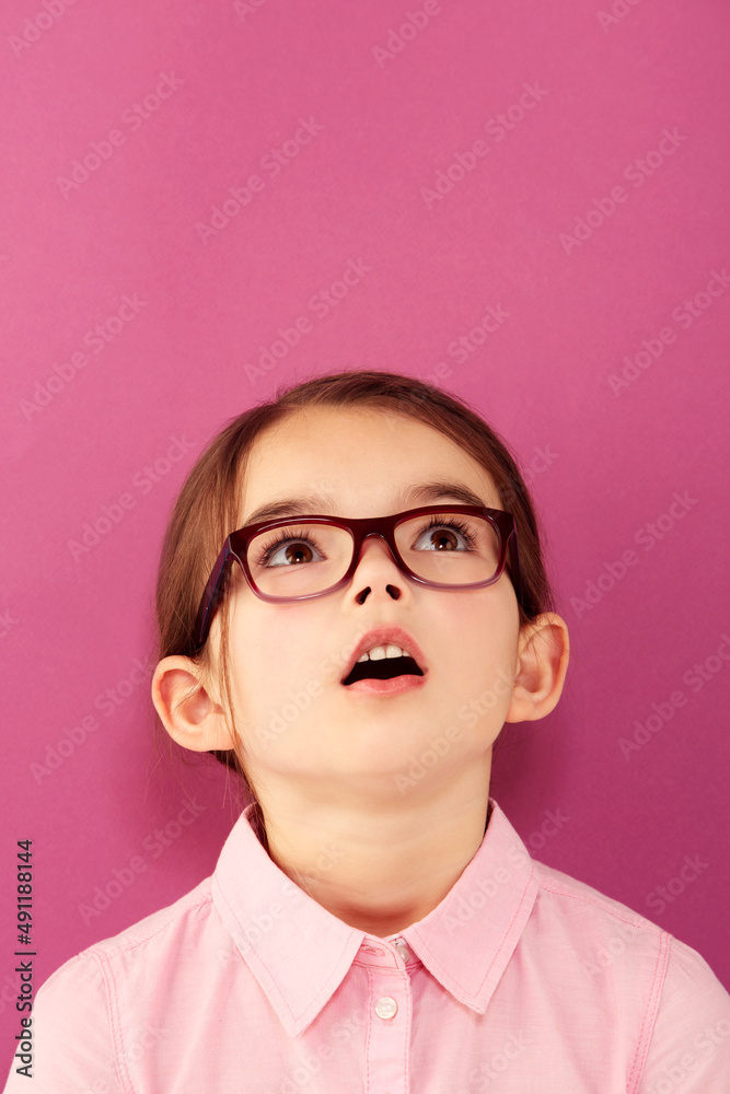 Childlike wonder. A little girl wearing spectacles looking up against a pink background.
