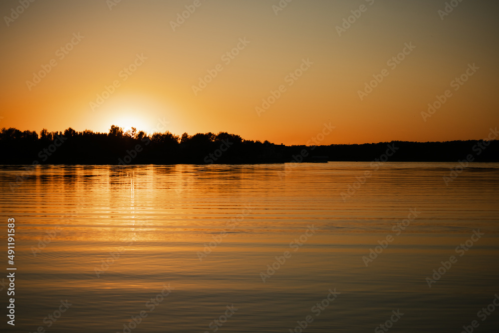 Calm rippled lake with sun almost set below horizon covering surface of water with its rays with trees on shore in background. Astonishing view full of yellow and orange colors.