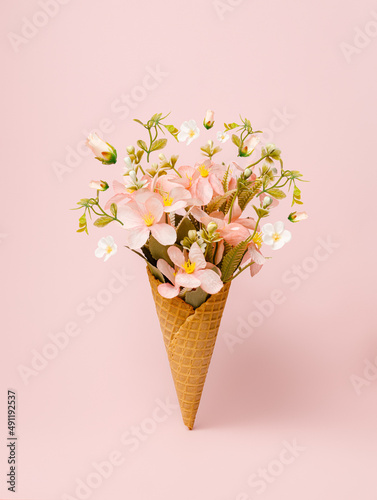 Ice cream cone filled with fresh flowers and leaves on pastel pink background. Creative floral spring bloom natural concept. Minimal summer dessert still life abstract idea.