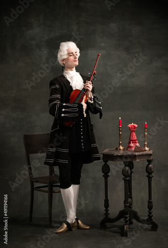 Portrait of young man wearing wig and vintage medieval outfit like famous composer playing violin isolated on dark green vintage background. photo