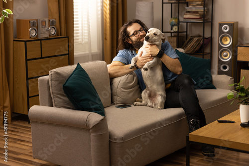 A bearded man in a blue denim shirt sits on the living room couch playing with his dog. The boy greets his pet after returning from work.