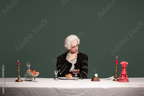 Portrait of young elegant man in peruke and vintage jacket sitting at table isolated on dark green background. Retro style, comparison of eras concept.