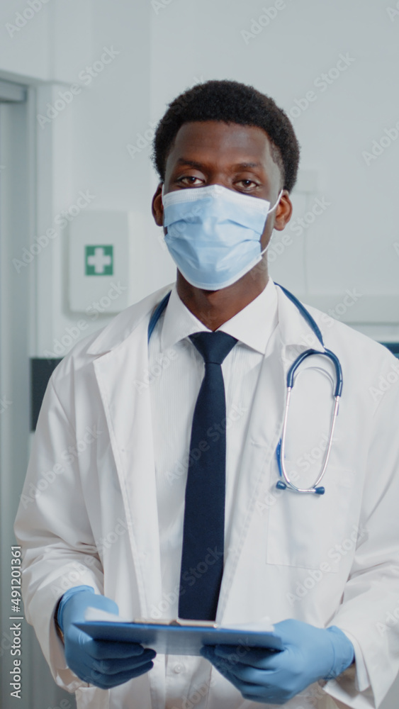 Portrait of man working as doctor standing in hospital ward, wearing face mask against coronavirus pandemic. Physician with stethoscope and white coat holding checkup documents. Portrait of man working as doctor standing in hospital ward, wearing face mask against coronavirus pandemic. Physician with stethoscope and white coat holding checkup documents.