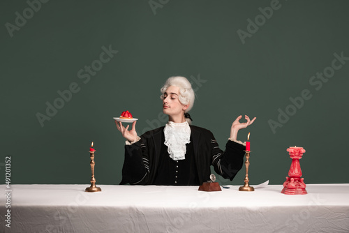 Portrait of young elegant man in peruke and vintage jacket sitting at table isolated on dark green background. Retro style, comparison of eras concept.