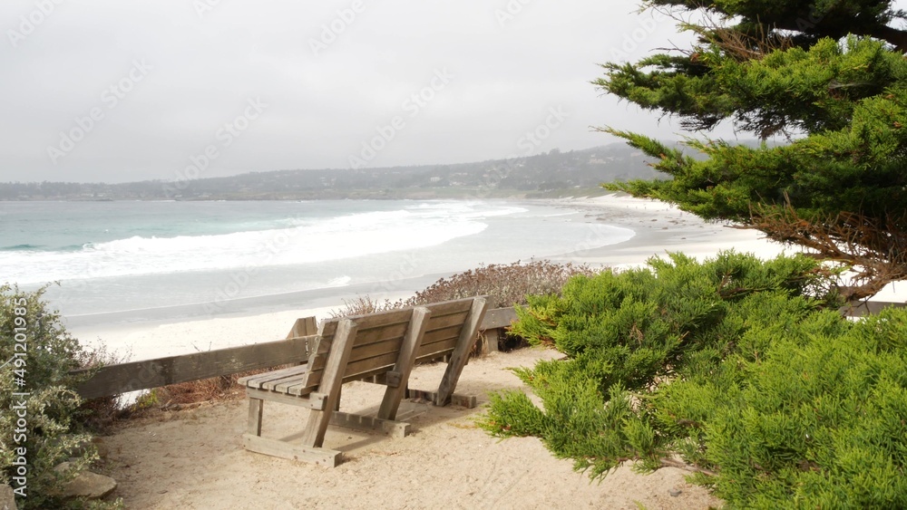 Empty wooden bench, rest on trail path, walkway or footpath. Carmel beach, Monterey ocean shore, California coast USA. Sea waves crashing. Waterfront beachfront pine cypress trees. Pathway or footway.
