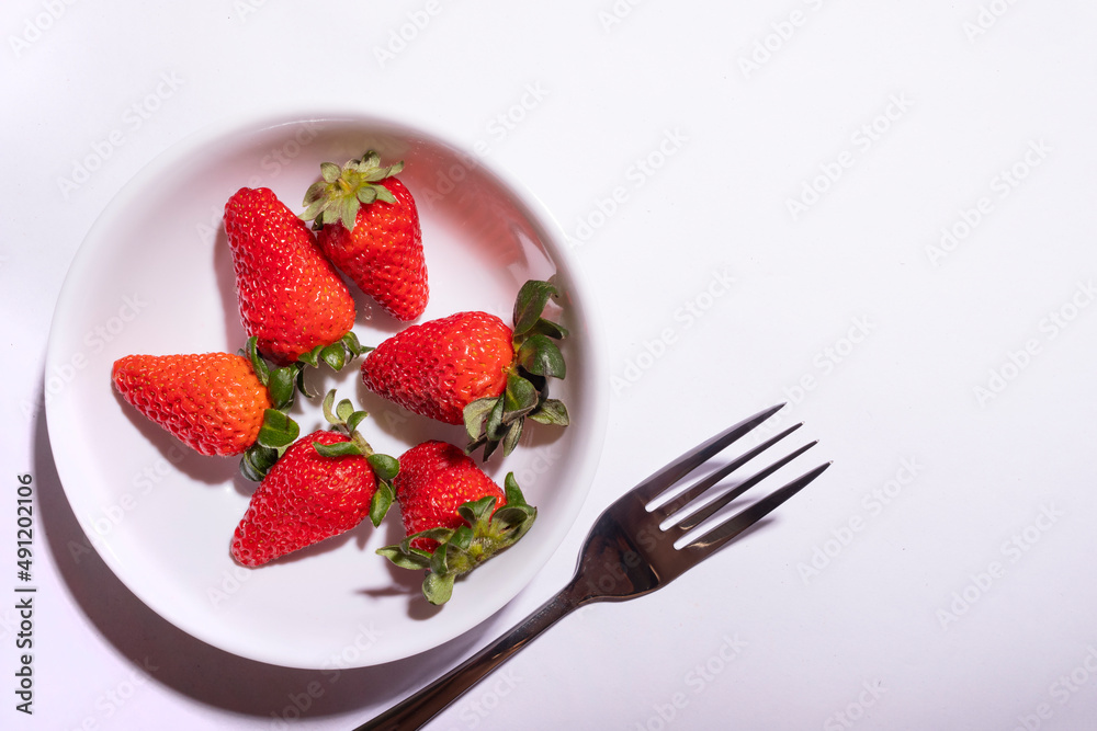 White bowl with red strawberries next to a fork. Perfect Breakfast with White Background and Empty Space for Text. White bowl with red strawberries next to a fork. Perfect Breakfast with White Background and Empty Space for Text.