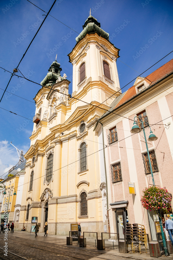 Obraz premium Linz, Austria, 27 August 2021: Baroque Ursuline Church of St. Michael with two towers, Narrow picturesque street with colorful buildings in historic center in medieval city at sunny summer day