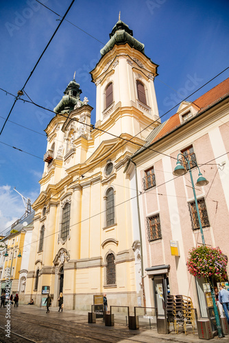 Linz  Austria  27 August 2021  Baroque Ursuline Church of St. Michael with two towers  Narrow picturesque street with colorful buildings in historic center in medieval city at sunny summer day
