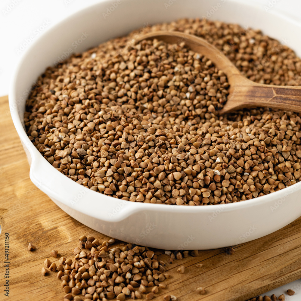 dry buckwheat groats in a white bowl on a wooden board with a wooden spoon, on a white background, close-up, square format dry buckwheat groats in a white bowl on a wooden board with a wooden spoon, on a white background, close-up, square format