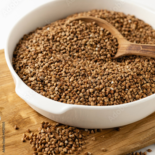 dry buckwheat groats in a white bowl on a wooden board with a wooden spoon, on a white background, close-up, square format