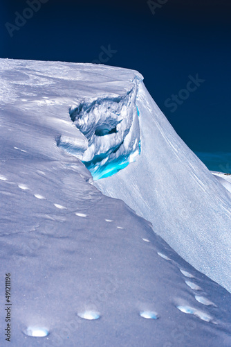 Shot of the high peek summit of a mountain with
a visible foot steps leading to a light blue i ce crack with a dark blue sky