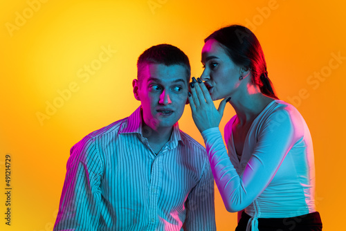 Portrait of two young people, woman whispering on man's ear, talking news isolated over orange background in neon lights