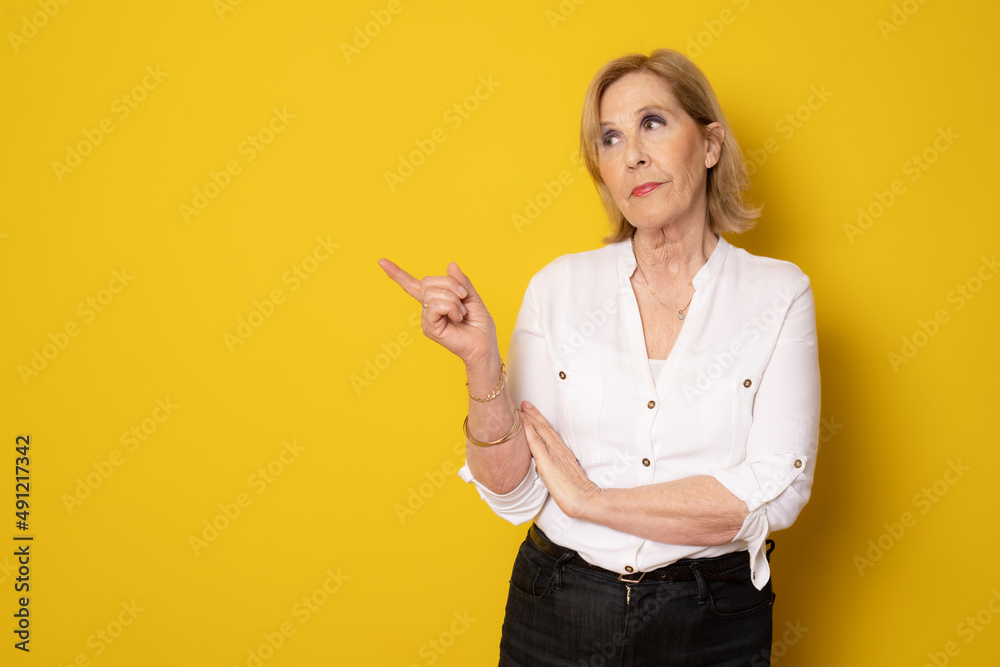 Smiling elderly blonde woman lady 70s years old in white casual shirt pointing index finger aside up on mock up copy space isolated on yellow background Smiling elderly blonde woman lady 70s years old in white casual shirt pointing index finger aside up on mock up copy space isolated on yellow background