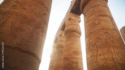 Massive pillars with intricate carved images and hieroglyphs in the Kamak Temple, Luxor, Egypt. 
Sun gleaming behind the pillars with a flare, slow sideways camera movement. photo