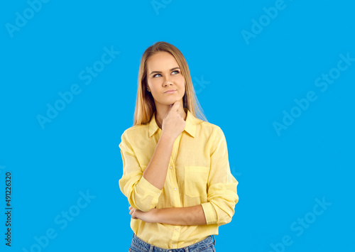 I wonder what it is. Curious young blonde woman holding hand on chin, looking away, thinking about something. Pretty girl in yellow shirt standing isolated on blue background, looking up and thinking