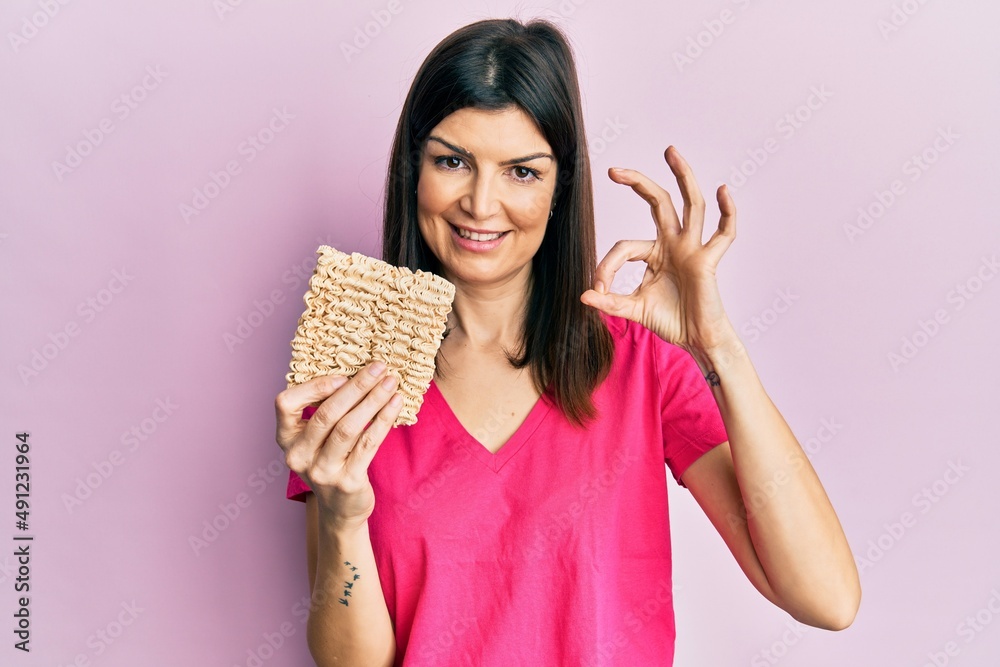 Young hispanic woman holding uncooked noodles doing ok sign with fingers, smiling friendly gesturing excellent symbol Young hispanic woman holding uncooked noodles doing ok sign with fingers, smiling friendly gesturing excellent symbol