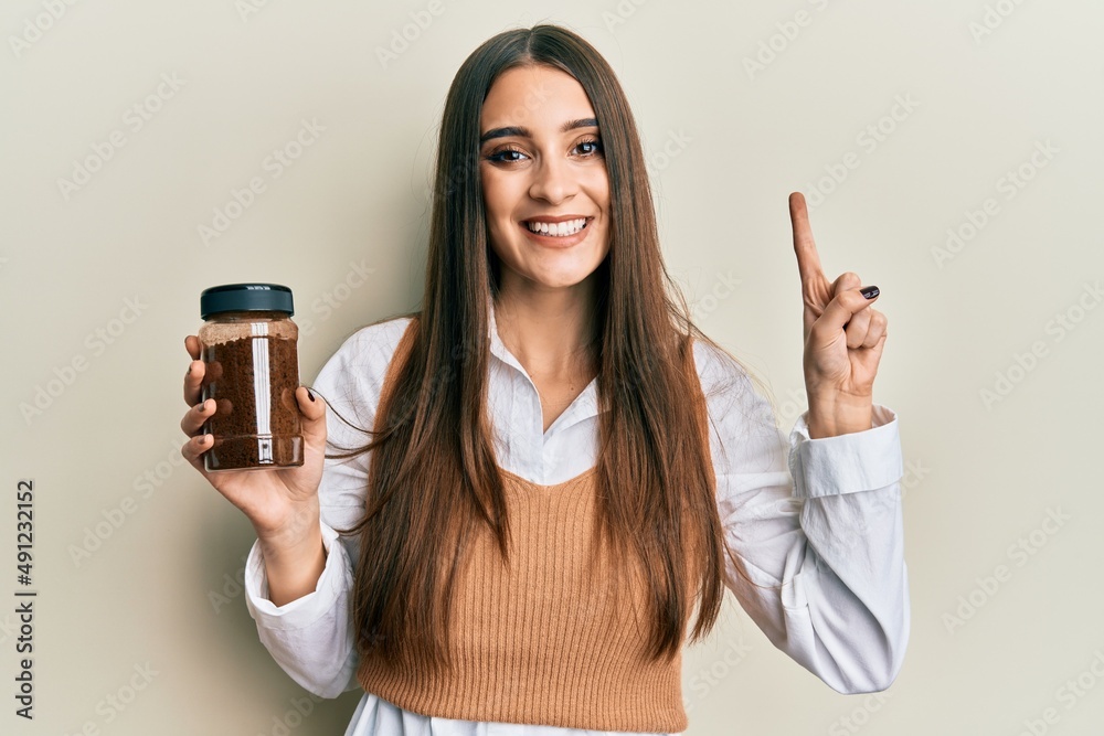 Beautiful brunette young woman holding soluble coffee smiling with an idea or question pointing finger with happy face, number one